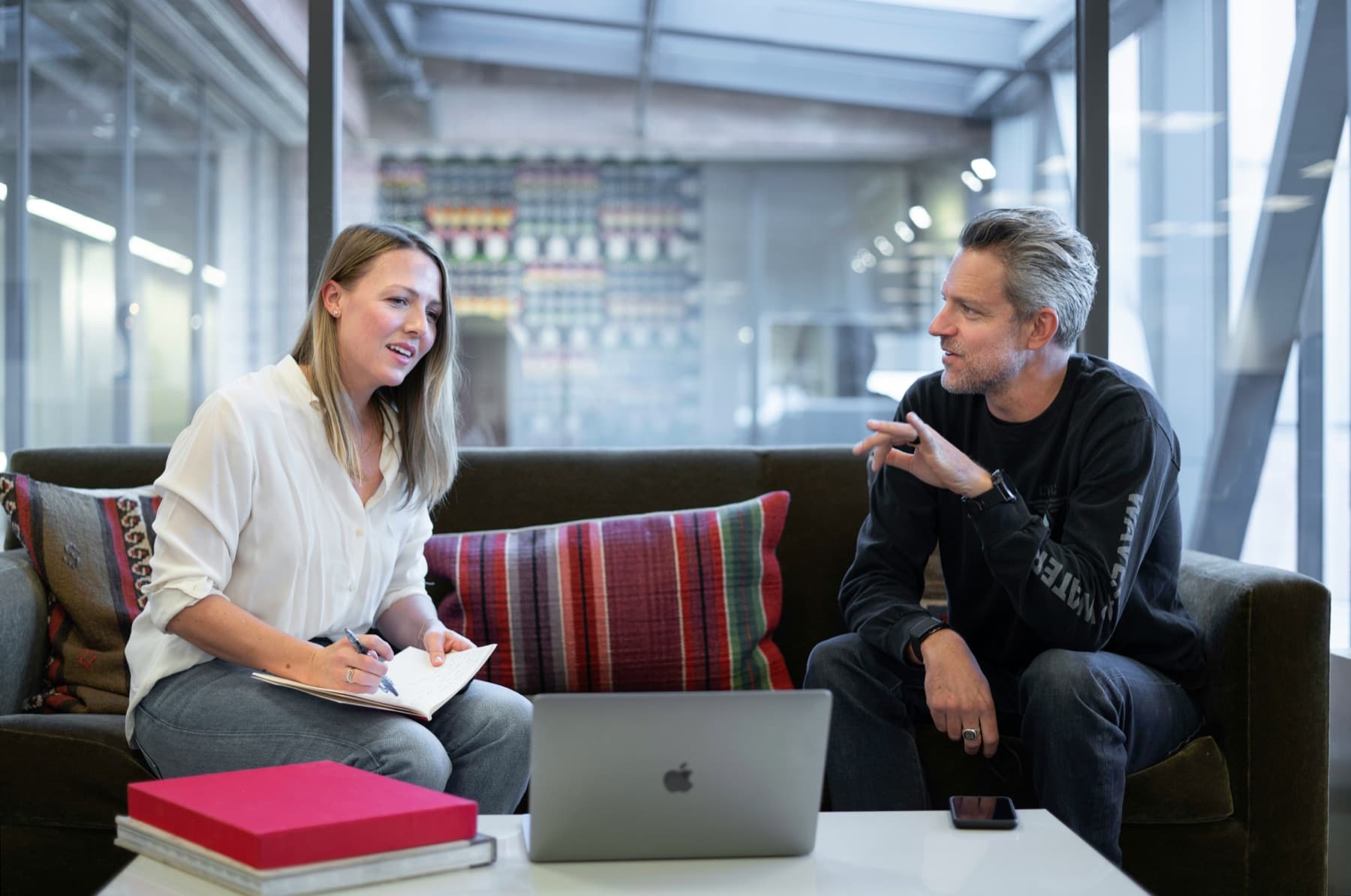 Two colleagues talking through notes on a sofa with a laptop between them